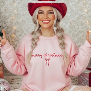 May include: A light pink sweatshirt with the text "merry christmas" in red script and a red bow graphic. The person is wearing a red cowboy hat with white trim and has blonde braids.