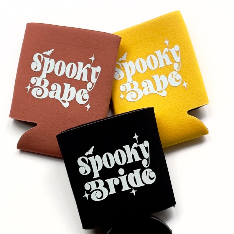 May include: Three different colored can coolers with the text "Spooky Babe" and "Spooky Bride" in white letters on a black, brown, and yellow background.