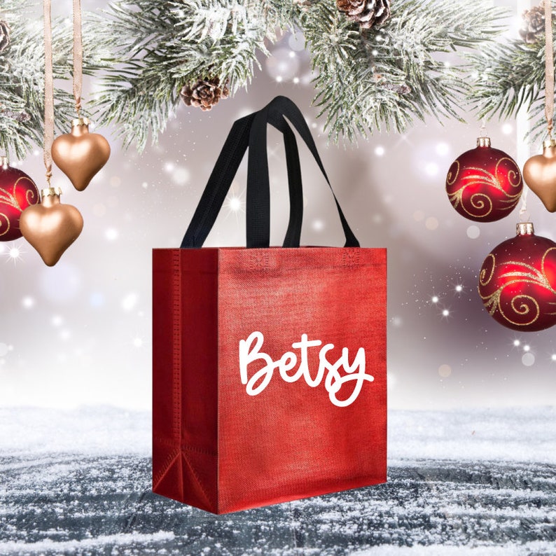 Op de afbeelding: Rode herbruikbare boodschappentas met zwarte handvatten en de naam "Betsy" in wit gedrukt op de voorkant. De tas is omringd door een besneeuwde achtergrond en kerstversiering.