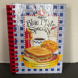 May include: A spiral-bound cookbook titled "Blue Plate Specials" with a red spine. The cover features illustrations of diner food, including a sandwich, onion rings, and a milkshake. The cover also has the text "Recipes your family & friends will love the most inspired by diners coast-to-coast."