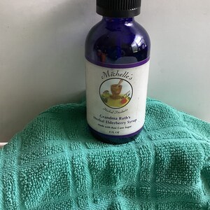 Könnte beinhalten: Eine dunkelblaue Glasflasche mit Michelle's Herbal Products Grandma Ruth's Herbal Elderberry Syrup. Das Etikett enthält eine Grafik von Mörser und Stößel. Die Flasche hat 118 ml und einen schwarzen Deckel. Die Flasche steht auf einem türkisfarbenen, strukturierten Handtuch.