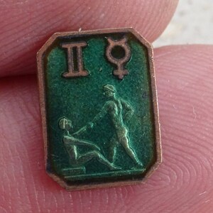 May include: A small, rectangular, vintage pin with a green enamel background and a bronze-colored border. The pin features the Gemini symbol, the Mercury symbol, and a depiction of two figures. The pin is held between two fingers.