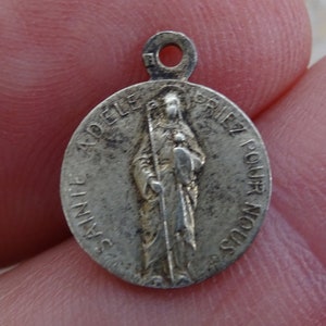 May include: Silver-toned metal pendant featuring a religious image of Saint Adele with the text "Sainte Adele Priez Pour Nous".