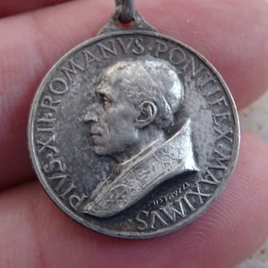 May include: Silver-toned metal pendant featuring a profile portrait of Pope Pius XII. The pendant is inscribed with the text "PIVS XII ROMANVS PONTIFEX MAXIMVS".