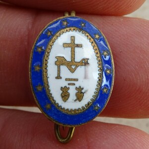 May include: Oval religious pendant with a blue border and gold accents. The center features a white background with a gold cross and other religious symbols. The pendant is held between two fingers.