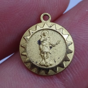 Puede incluir: Pendiente de metal dorado con un diseño de rayos de sol y una imagen en relieve de un santo sosteniendo una rama.