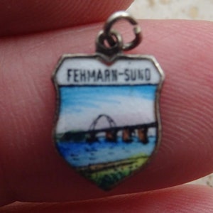 May include: A silver charm in the shape of a shield with a blue, white, and green enamel design. The charm features a bridge and the text "FEHMARN-SUND".