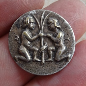 May include: A round, silver-toned metal medallion featuring a relief of two kneeling figures holding a central stalk. The figures, with flowing hair, are depicted in a classical style. The medallion has a textured surface.