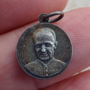 Op de afbeelding: Zilverkleurige ronde hanger met een reliëfbeeld van een mannengezicht. De hanger heeft bovenaan een lus om aan een ketting te bevestigen.