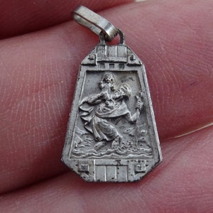 May include: Silver pendant featuring a detailed image of Saint Christopher carrying a child. The pendant is in the shape of a shield with a decorative border.