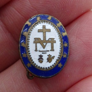 May include: A gold-colored oval pin with a blue enamel border and white enamel center. The center features a gold cross, a gold 'M' with a gold crown above it, and a gold dove with a gold heart below it.
