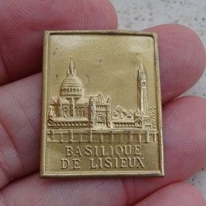 Op de afbeelding: Een gouden metalen pin met een reliëfbeeld van een kerkgebouw en de tekst "BASILIQUE DE LISIEUX".