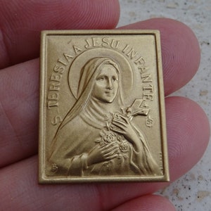 Op de afbeelding: Een gouden metalen religieuze medaille met een reliëfbeeld van een vrouw in een witte jurk die een bos rozen vasthoudt. De tekst "S. TERESIA JESU INFANTE" is rond de afbeelding gegraveerd.
