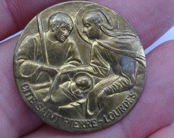May include: A gold-colored metal religious medal depicting the nativity scene with the text "CITE SAINT PIERRE - LOURDES".