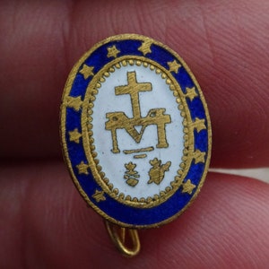May include: A gold-colored oval pin with a blue enamel border and white enamel center. The center features a gold-colored cross, a stylized 'M' with a cross above it, and two small gold-colored symbols below. The border is decorated with gold-colored stars.
