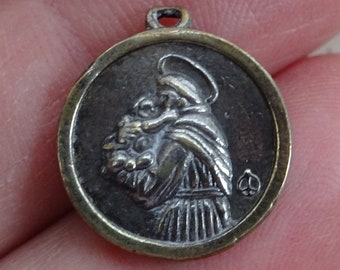 May include: A round, antique-style pendant with a detailed relief of a figure holding flowers. The pendant has a dark silver-tone center with a gold-tone border and a small loop at the top for a chain.