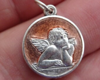May include: A circular pendant featuring a silver cherub design set against a copper-toned backdrop. The cherub, complete with wings, rests its head on its hand. The pendant is framed by a silver border and has a small loop.
