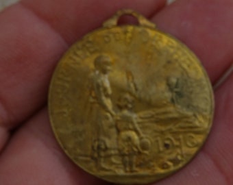 Könnte beinhalten: Ein goldfarbener Metallanhänger mit einem Reliefbild einer Frau und eines Kindes. Der Anhänger trägt den Text "Mothers of the World" und "1916" am Rand.