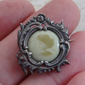 May include: A silver cameo brooch with an ornate, scrolled frame and a pale green cameo of a woman's profile.