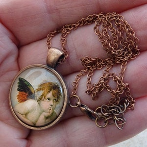 May include: A copper chain necklace with a round pendant featuring a vintage image of a cherub with wings. The pendant has a glass dome and a bronze frame.