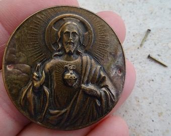 May include: A round, bronze-colored metal medallion with a raised image of Jesus Christ. He is wearing a robe and has a heart with thorns on his chest. The medallion has a halo around his head and a sunburst design behind him.