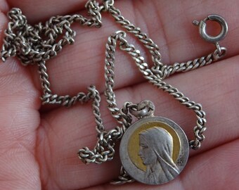 May include: A silver chain necklace with a round pendant featuring a gold-toned image of the Virgin Mary.