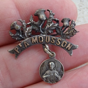 May include: Silver and gold tone religious pin with a depiction of Jesus Christ and the words "PTA MOUSSON".