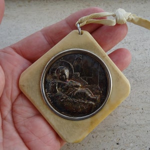 May include: A white square ornament with a silver rimmed circular image of baby Jesus in a manger. The ornament has a cream colored ribbon attached.