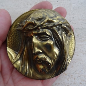May include: A gold-toned metal plaque with a raised image of Jesus Christ wearing a crown of thorns. The plaque has a textured background.