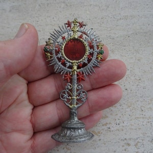 May include: A silver-toned religious monstrance with a red center and ornate detailing. The sunburst design features red, green, and gold accents. The base is detailed and the piece is held in a hand.