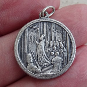 May include: Silver pendant featuring a religious scene with a figure in a robe holding a cross, surrounded by other figures in robes. The pendant is circular and has a raised design.