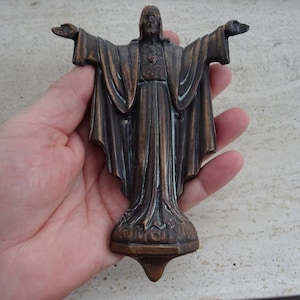 May include: Bronze statue of Jesus with arms outstretched. The figure is in flowing robes, with a detailed face and a heart emblem on the chest. The statue has a decorative base and is held in a hand.
