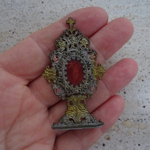 May include: A small, ornate, silver-toned metal religious reliquary with a red glass center and gold accents. The reliquary is in the shape of a cross and has a base.