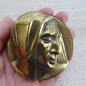 May include: A gold-toned metal plaque with a raised relief sculpture of a woman's face. The woman is wearing a head covering and her eyes are closed. The plaque has a textured background.