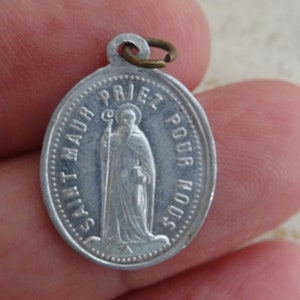 May include: Silver oval-shaped pendant with a religious image of a saint holding a staff and a book. The pendant has the text "SAINT MAUR PRIEZ POUR NOUS" around the edge.