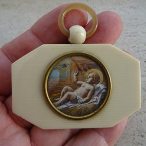 May include: A small, ivory-colored pendant with a gold-toned frame. The frame encloses a miniature painting of the baby Jesus in a manger. The pendant has a loop at the top for hanging.