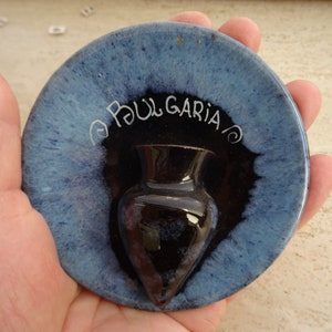 Può includere: Un piatto in ceramica blu e nero con un piccolo vaso nero al centro. Il piatto ha la scritta "BULGARIA" in bianco.