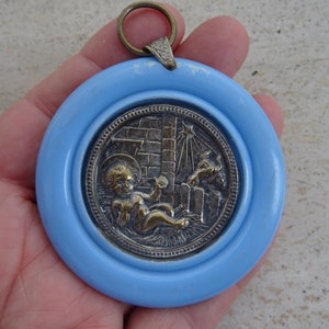 May include: A blue plastic framed pendant with a silver and gold metal medallion depicting the nativity scene. The medallion features a baby Jesus, a star, and an ox.
