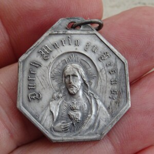 May include: Silver octagonal pendant with a depiction of Jesus Christ with a heart and a crown of thorns. The pendant has the text "Durch Maria zu Jesus" around the edge.
