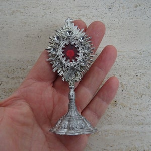 May include: A silver-toned metal monstrance with a red glass center and a decorative base. The monstrance is adorned with rhinestones.