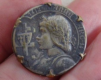 Könnte beinhalten: Eine silberfarbene Metallmünze mit dem Profil einer Frau, die eine Blätterkrone trägt. Die Münze trägt den Text "1412 J. ANNA ARC. 14" am Rand.