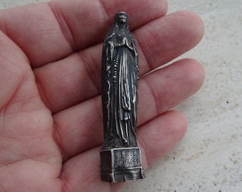 May include: A silver-colored statue of the Virgin Mary, standing on a base with the inscription "N.D. de Lourdes".