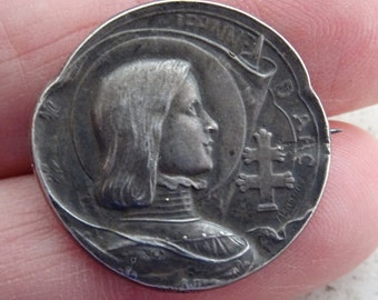 Könnte beinhalten: Silberfarbener Metallstift mit einem Profil einer Frau, die einen Helm trägt, und einem Kreuz mit dem Text "Jeanne D'Arc" am Rand.