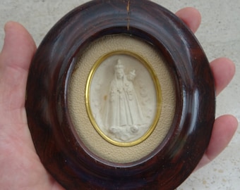 May include: A small oval-shaped framed image of a white ceramic sculpture of a woman in a white dress with a crown on her head. The frame is made of dark wood with a gold trim.