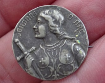 Könnte beinhalten: Eine silberfarbene Metallstiftnadel mit einer Darstellung der Jeanne d'Arc, die ein Schwert hält. Die Nadel trägt die Aufschrift "JOHANNA D ARC".