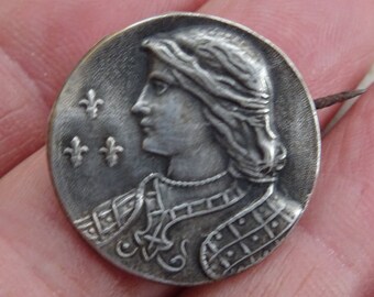 Könnte beinhalten: Silberfarbener Metallstift mit dem Profil einer Person, die eine Krone und eine Rüstung trägt. Im Hintergrund sind drei Lilien zu sehen.