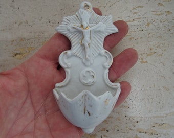 Puede incluir: Fuente de agua bendita de porcelana blanca con un diseño pintado en oro. La fuente presenta una imagen en relieve de un crucifijo con una figura de Jesucristo.