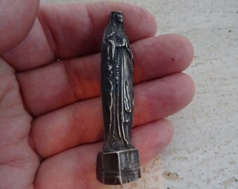 May include: A small silver statue of the Virgin Mary, standing on a square base. The statue is detailed and shows the Virgin Mary with her hands clasped in prayer.