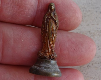 May include: A small, gold-colored statue of the Virgin Mary. The statue is standing on a round base and is wearing a long robe. The statue is about 3 inches tall.