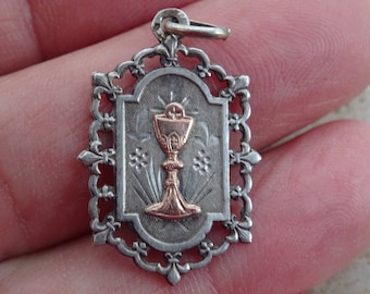 May include: Silver and gold religious pendant with a chalice design. The chalice is gold and the pendant is silver with an ornate border.